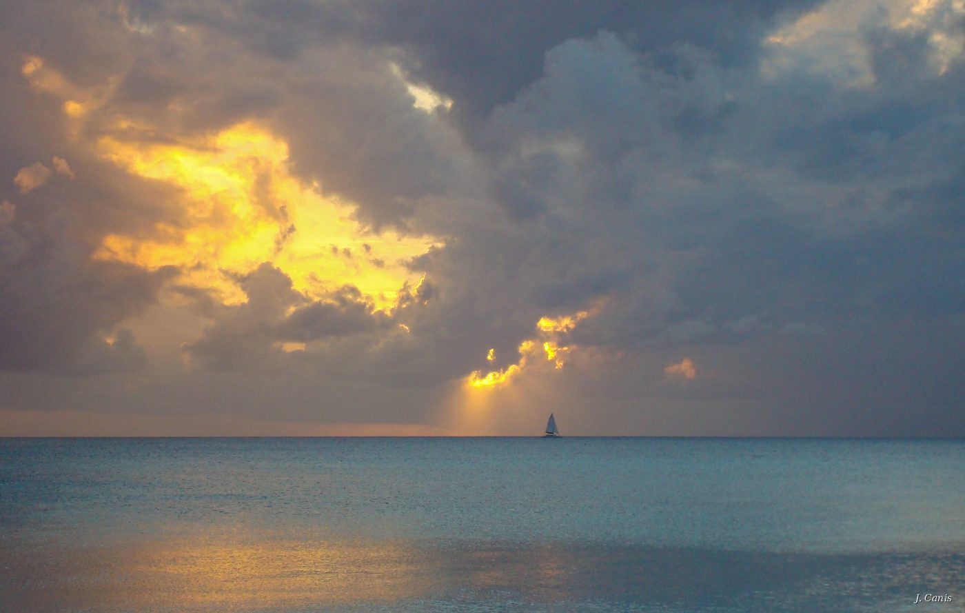 Smooth Sailing — lone sailboat beneath dramatic backlit storm clouds over Caribbean Sea, Cayman Islands photography by Jon Canis