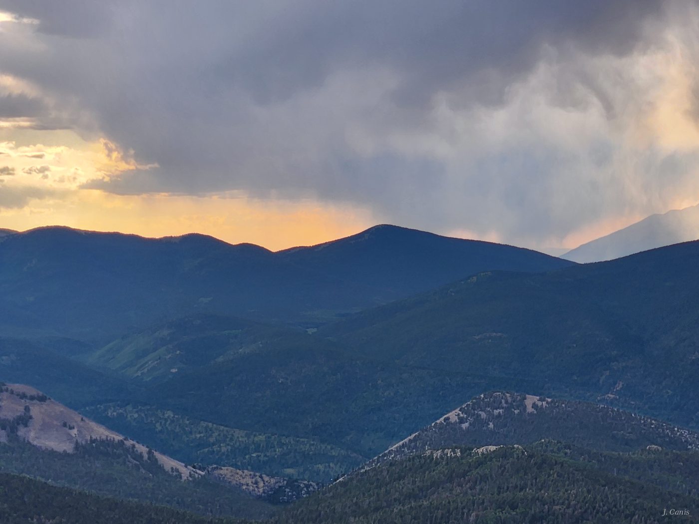 Mountain Storm — layered blue Colorado ridges under dramatic storm clouds with warm sunset light, photography by Jon Canis